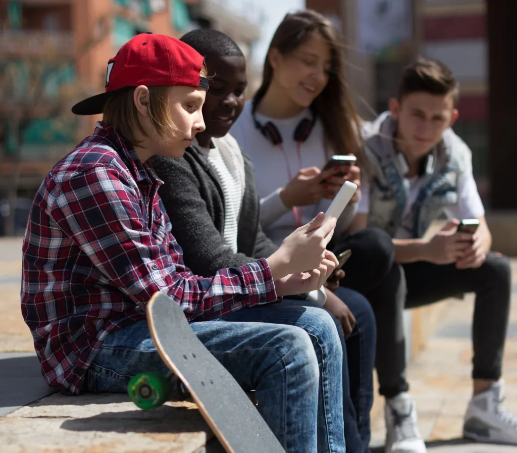 A group of young friends sitting on a wall looking at each other's phone screens. A skateboard rests on the wall. 