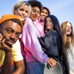 A diverse group of seven young friends smiling and posing together outdoors against a blue sky, one holding a skateboard