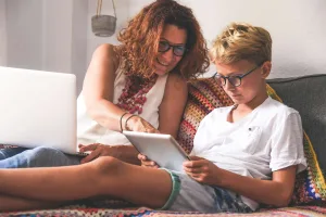A mother and her young son sitting on the couch together, looking at something on a tablet