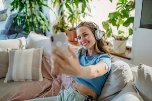 A smiling young woman relaxing on a couch with headphones, taking a selfie, wearing a continuous glucose monitor on her arm and an insulin pump at her waist