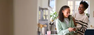 A mother and teenage daughter laughing together in a bright kitchen, the teen holding a smartphone while the mother rests her hand on her daughter's shoulder.