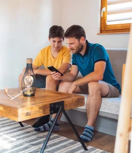 A father and teenage son sitting together on a couch, both looking at a smartphone, with a wooden coffee table and decorative lamp in front of them.