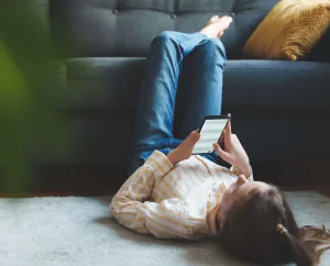 A young woman lying on the floor with her legs up on a couch, scrolling on her phone